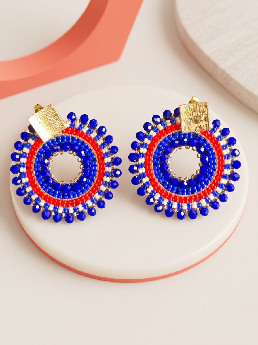 Aros circulares artesanales tejidos a mano en color azul, rojo y blanco con cristales y base bañada en oro, presentados sobre una superficie minimalista.