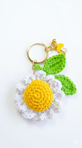 Llavero de margarita tejida a mano con centro amarillo y pétalos blancos, incluye hojas verdes tejidas, un dije de hongo amarillo y herrajes dorados.